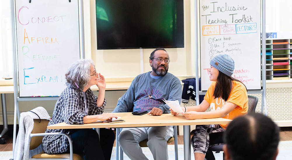 Three actors sitting around a table with white boards charts behind them